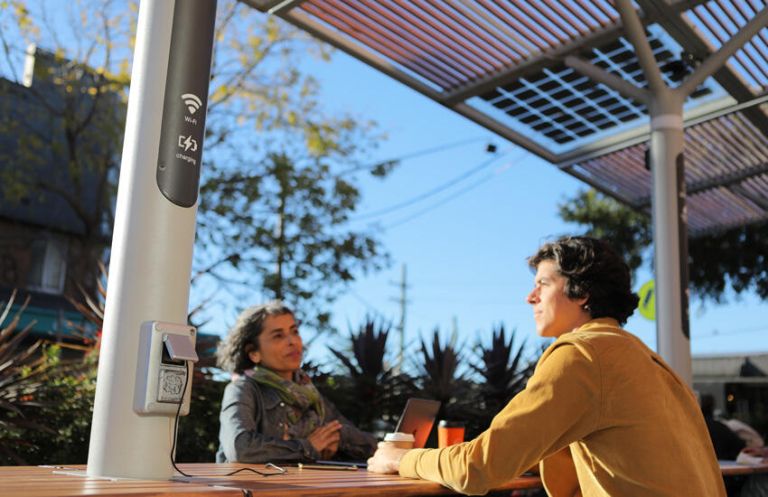 Two people using wi-fi and a charging station at an outdoor workstation known as a 'chillout hub'.