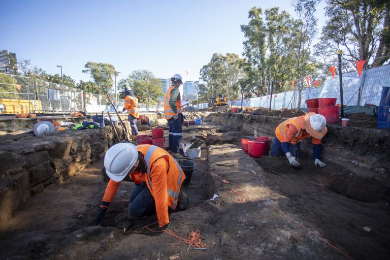 Several workers in high-visibility clothing are on the ground of a dig-site in the shade, conducting excavations with their hands and some tools.