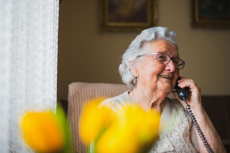 Silver-haired senior wearing glasses and a bright yellow top talking into handset.
