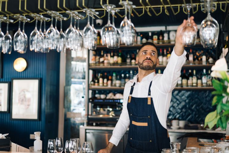A bartender in a white shirt and blue apron, hanging wine glasses