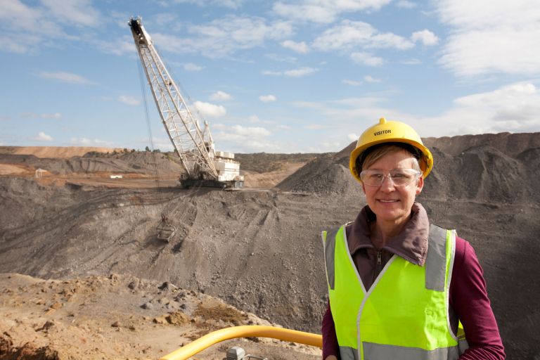 A person in a helmet a hi-vis vest stands with a coal mine in the background