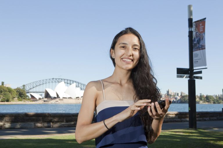 A person holding a mobile phone in front of the Sydney Harbour Bridge and Opera House in Sydney, NSW.