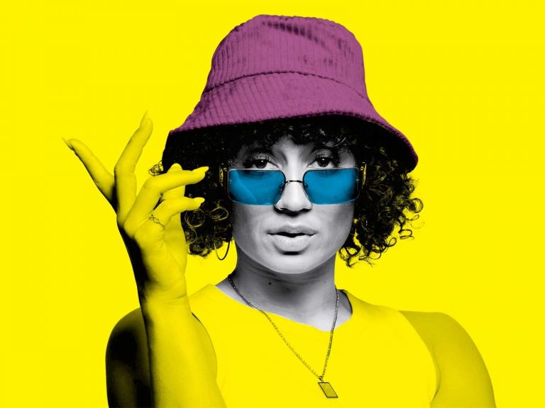 A woman in a bucket hat and sung glasses stares to camera with her hand posed next to her face.