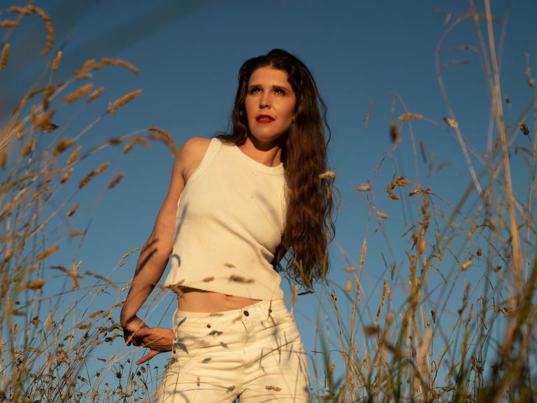 Woman dressed in white singlet & white jeans, blue sky background, long, dry grass foreground.