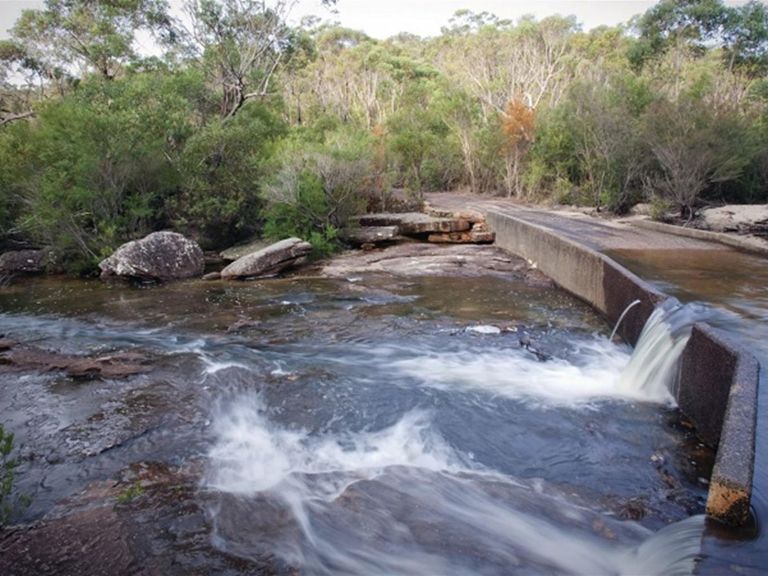 Water flows over concrete Battery Causeway against a backdrop of bushland in Heathcote National Park