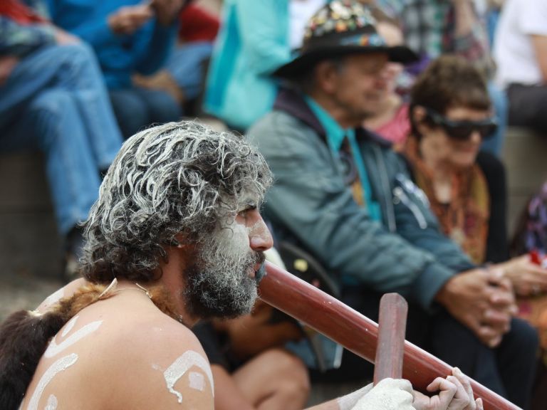 An Australian Aboriginal man wearing facepaint plays the didgeridoo for a crowd.