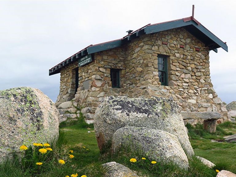 View of historic stone hut set in alpine grassland, with large boulders and wildflowers in the