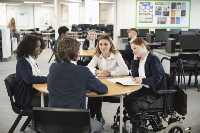 Four students around a table in navy and white school uniform - 1 Black and 3 white mixed gender, discussing notes.