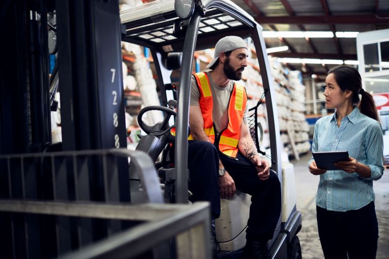 A man on a forklift speaks to a colleague in a warehouse