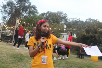 A female volunteer with medium length brown curly hair is holding a microphone in her right hand and a script in her out-streched left hand. She is wearing a red bandana over her hair and a yellow shirt reading Bring It On! Festival