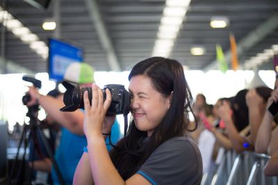 A young female photographer with long dark hair and a grey volunteer shirt is taking a photo at a youth festival with a large Canon camera