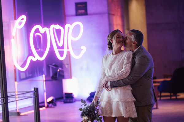 A bride and groom share an embrace next to a neon pink sign that says love.