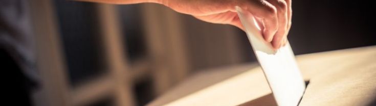 A person's hand holding a vote over a ballot box.