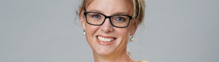 Stephanie Cooke smiling at the camera wearing glasses and a yellow blouse in front of a grey background.