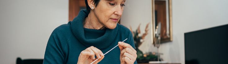 A woman in a green sweater sits at home studying the instructions of a rapid antigen test with a long nasal swab in hand