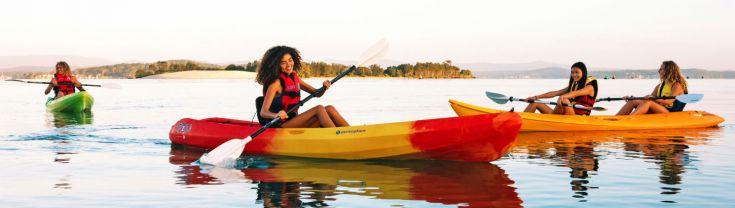Four young people sit in three yellow, orange and green canoes on a blue lake. They each wear and orange life jacket and hold oars