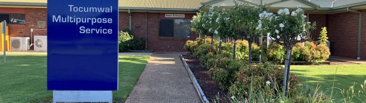 Main entrance to the Tocumwal Multipurpose Service. A walkway leads to the main entrance. To the left of the image, a blue sign with the NSW state logo shows the name of the service and "Murrumbidgee Local Health District". A landscaped green space sits to the right of the walkway. 