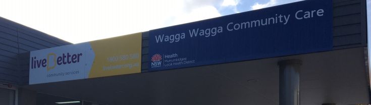 Main entrance to the Wagga Wagga Community Care centre. There is a bicycle ramp for 4 bicycles outside the main entrance. To the right, there are a couple of car parking spaces. Along the top left of the building's entrance awning the centre's signage shows 'live Better community living' and 'Wagga Wagga Community Care'.  