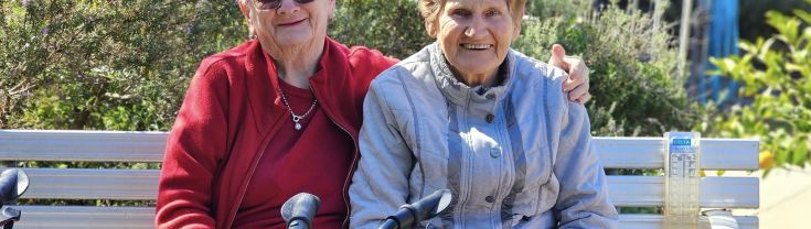 An older woman wearing a red jacket sits with her left arm around another older woman on a park bench. They are facing forward and smiling. A walking aid is in front of them. 