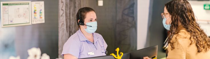 A hospital receptionist with a face mask speaks to a visitor who is also wearing a face mask at a hospital's front desk.
