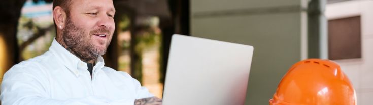 A man wearing a white shirt sitting at a table using his laptop with an orange construction hat next to it