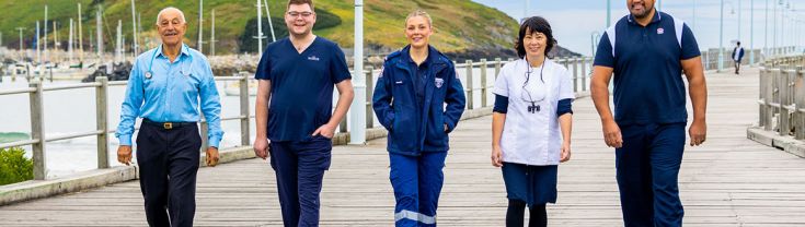 5 essential workers walking across a timber bridge in Coffs Harbour with small boats, a hill and blue sky with streaky clouds in the background
