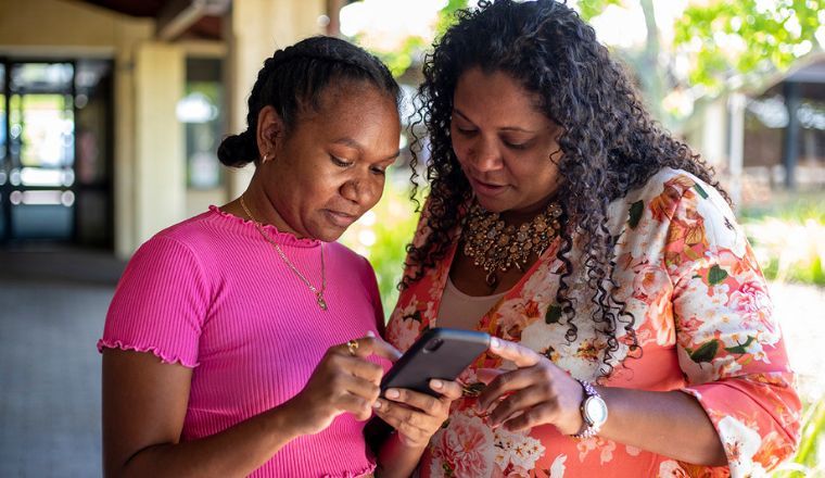 Two Indigenous women wearing brightly coloured tops standing outside looking at an iPhone
