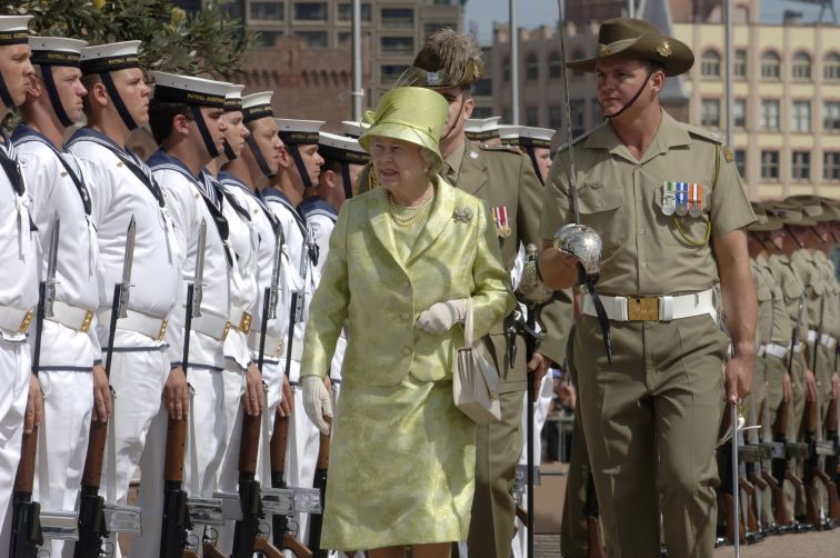 Her Majesty Queen Elizabeth II is walking with Navy officials inspecting the Australian Navy. Her Majesty is wearing a light lime green skirt and jacket with a matching hat. The official is wearing a khaki green uniform and the Navy Members are wearing their white uniforms.