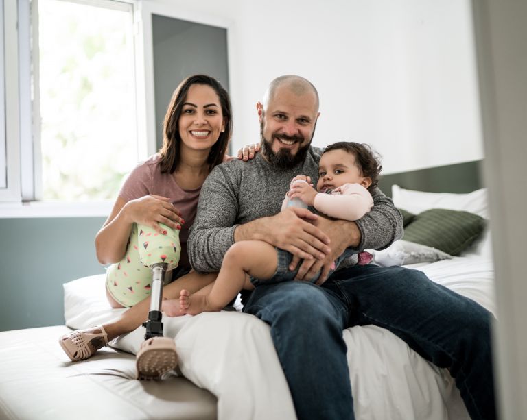 A family of three. A mother, father and infant girl, sitting on a bed, smiling.
