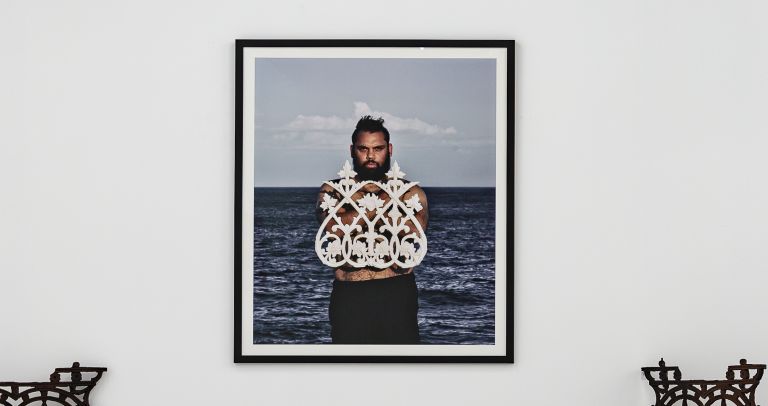 A photo hung on a white wall of a first nations man with a beard, the sea behind him holding a white sculpture over his bare chest, staring directly at the camera. 
