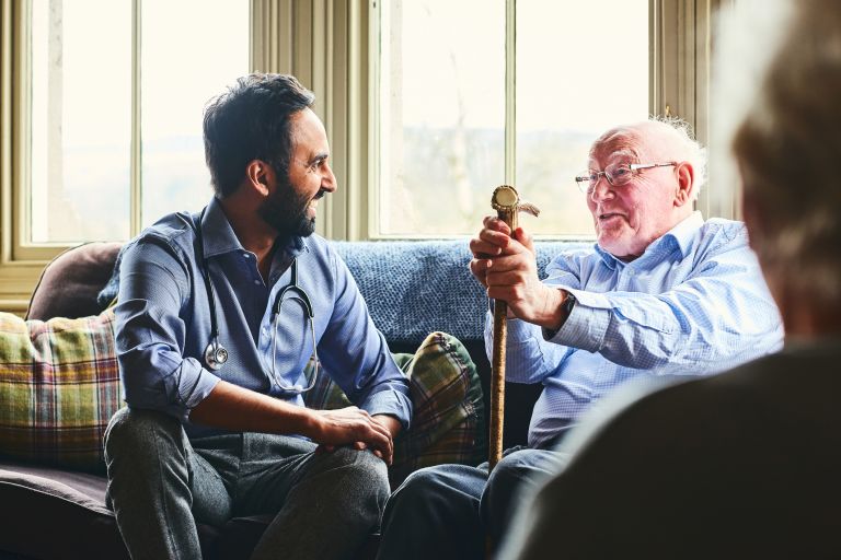 An elderly man and a doctor are sitting on a couch in an aged care facility talking. Both men look happy. 