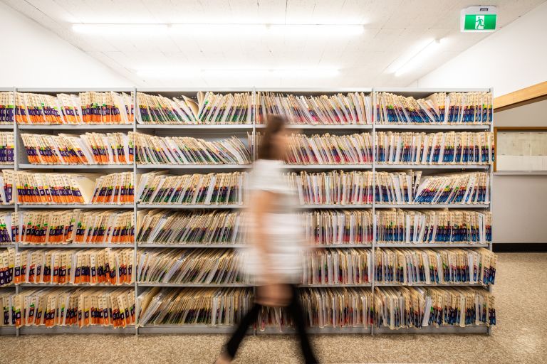 A blurry image of a woman walking past a shelf with files in the background.