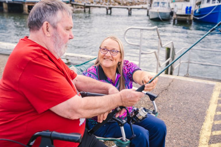 A man wearing a red shirt holds a fishing rod and sits next to a woman smiling and looking toward him while helping to hold the fishing rod.